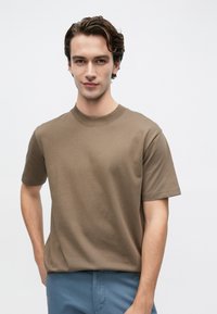 Καφέ κοντομάνικη μπλούζα από βαμβάκι. Λαιμόκοψη τύπου crew με άνετη εφαρμογή. Συνδυάζεται με μπλε παντελόνι, το μοντέλο στέκεται μπροστά σε απλό φόντο.