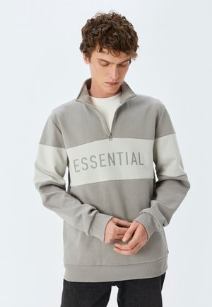 Grå sweatshirt med en kvart-zip krave, prydet med et fedt "ESSENTIAL" print i lysere grå over brystet. Blødt stof, afslappet pasform.