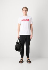 Bílý tričko s velk�ým červeným nápisem "HUGO", v kombinaci s černými cargo kalhotami, černými skládacími sandály a černou kabelkou přes rameno. Jednoduchý design.