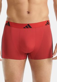 Mužský torso oblečený v červených boxerkách Adidas s černým logem na pásku a jemným tečkovaným vzorem na bocích, stojící na bílém pozadí.