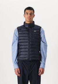 Marineblauwe gewatteerde vest met hoge kraag, rits aan de voorkant en horizontale quilt. Heeft een klein groen logo op de borst. Gelaagd over een gestreept shirt.