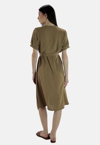 Brun midi-klänning med korta uppvikta ärmar, bälte i midjan, mjuk textur och sidoflikar, bärs med platta sandaler. Bakvy visas.
