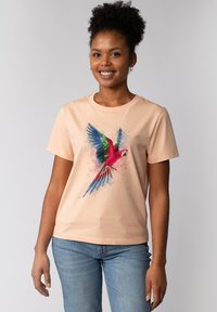 Perzikkleurig katoenen t-shirt met een kleurrafiek van een papegaai met blauwe, groene en rode accenten, tegen een spetter achtergrond.