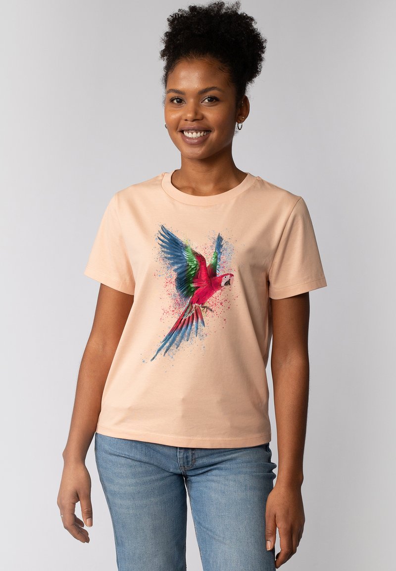Perzikkleurig katoenen t-shirt met een kleurrafiek van een papegaai met blauwe, groene en rode accenten, tegen een spetter achtergrond.