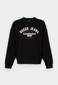 Felpa nera in cotone, con colletto tondo e spalle scese. Presenta la scritta "GUESS JEANS" e le date in un testo ricamato a contrasto.