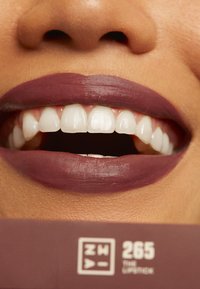 Matte deep plum lipstick on full lips, showcasing bright white teeth and smooth texture, with a color label displaying "265 THE LIPSTICK."