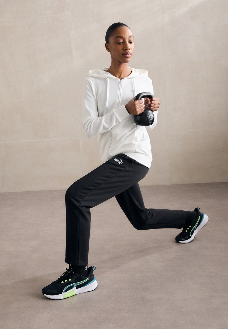 Fehér cipzáras kapucnis pulóver és fekete nadrág; fekete kettlebellt tart. A cipők feketék zöld részletekkel. Simább, sportos anyagok minimális dizájnnal.