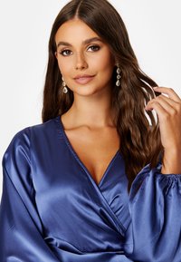 Szatynowa, głęboko granatowa bluzka z dekoltem w kształcie V i bufkami. Charakteryzuje się wyraźnym blaskiem i miękką teksturą; noszona z długimi, łezkowymi kolczykami.