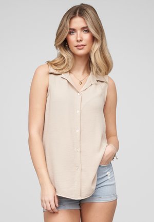 Kvinde med bølget blondt hår iført en ærmeløs beige knappet top og lyseblå cowboyshorts, stående med den ene hånd i lommen.