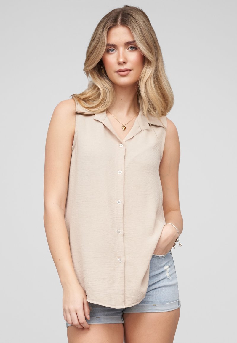 Femme aux cheveux blonds ondulés portant un haut beige sans manches boutonné et un short en jean bleu clair, debout avec une main dans sa poche.