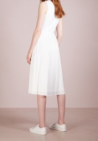 Bílá midi šaty s plisováním a bezrukávovým designem, obuté do bílých tenisek. Tkanina vypadá hladce s vertikální texturou.