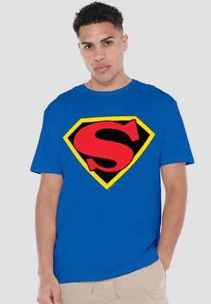 Young man with short curly hair wearing a blue T-shirt with a large red "S" inside a yellow-bordered black diamond shape on the chest.