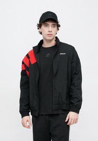 Μαύρο ελαφρύ μπουφάν με κόκκινες διαγώνιες ρίγες στο αριστερό μανίκι, με ψηλό γιακά και κλείσιμο με φερμουάρ. Συνδυασμένο με μαύρο μπλουζάκι.