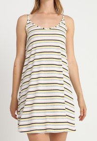 Mujer vistiendo un vestido sin mangas, de longitud hasta la rodilla, con rayas horizontales en blanco, negro, amarillo y rosa sobre un fondo claro.