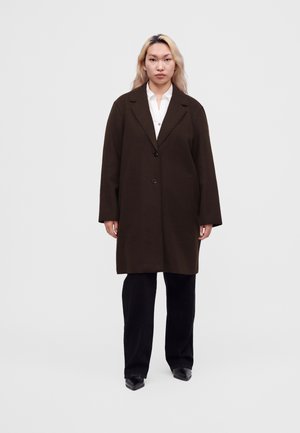 Personne aux longs cheveux blonds portant un manteau oversize marron foncé, une chemise blanche, un pantalon noir et des chaussures noires, debout devant un fond uni.