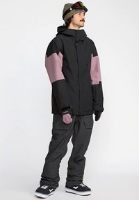 Chaqueta de nieve negra y rosa con capucha, con cierre de cremallera en el frente, grandes bolsillos y puños elásticos, combinada con pantalones de nieve grises y botas negras.