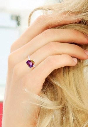 Zilveren ring met een hartvormige paarse edelsteen met groene accenten, gezet op een gladde band, weergegeven op een hand.