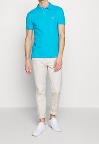 Tyrkysové polo tričko s krátkým rukávem a límečkem, s vyšitým logem, v kombinaci se světle béžovými chinos kalhotami s ohrnutými nohavicemi a bílými teniskami.