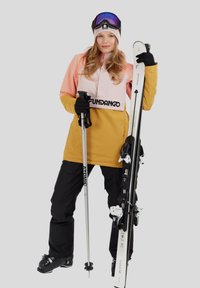 Woman in pink and mustard ski jacket wearing black pants, gloves, and goggles holds skis and a ski pole against a plain gray background.