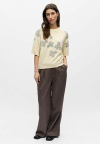 Pull crème à manches courtes avec motifs de papillons brodés. Associé à un pantalon large marron foncé et des chaussures noires. Textures lisses.