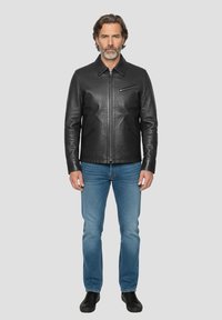 Schwarze Lederjacke mit spitzem Kragen, Frontreißverschluss und Reißverschlusstasche auf der Brust. Getragen über blauen Jeans und schwarzen Schuhen.