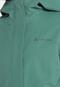 Veste imperméable verte avec capuche et fermeture éclair frontale, avec logo VAUDE sur la poitrine.