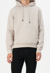 Hoodie beige chiaro realizzato in tessuto morbido, con tasca frontale, lacci regolabili e polsini e orlo a coste. Indossato con jeans neri.