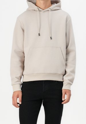 Hoodie beige chiaro realizzato in tessuto morbido, con tasca frontale, lacci regolabili e polsini e orlo a coste. Indossato con jeans neri.