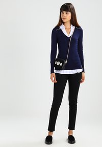 Navy fitted sweater with zip accents, layered over white collared shirt; paired with black slim pants and a small black handbag with floral detail.