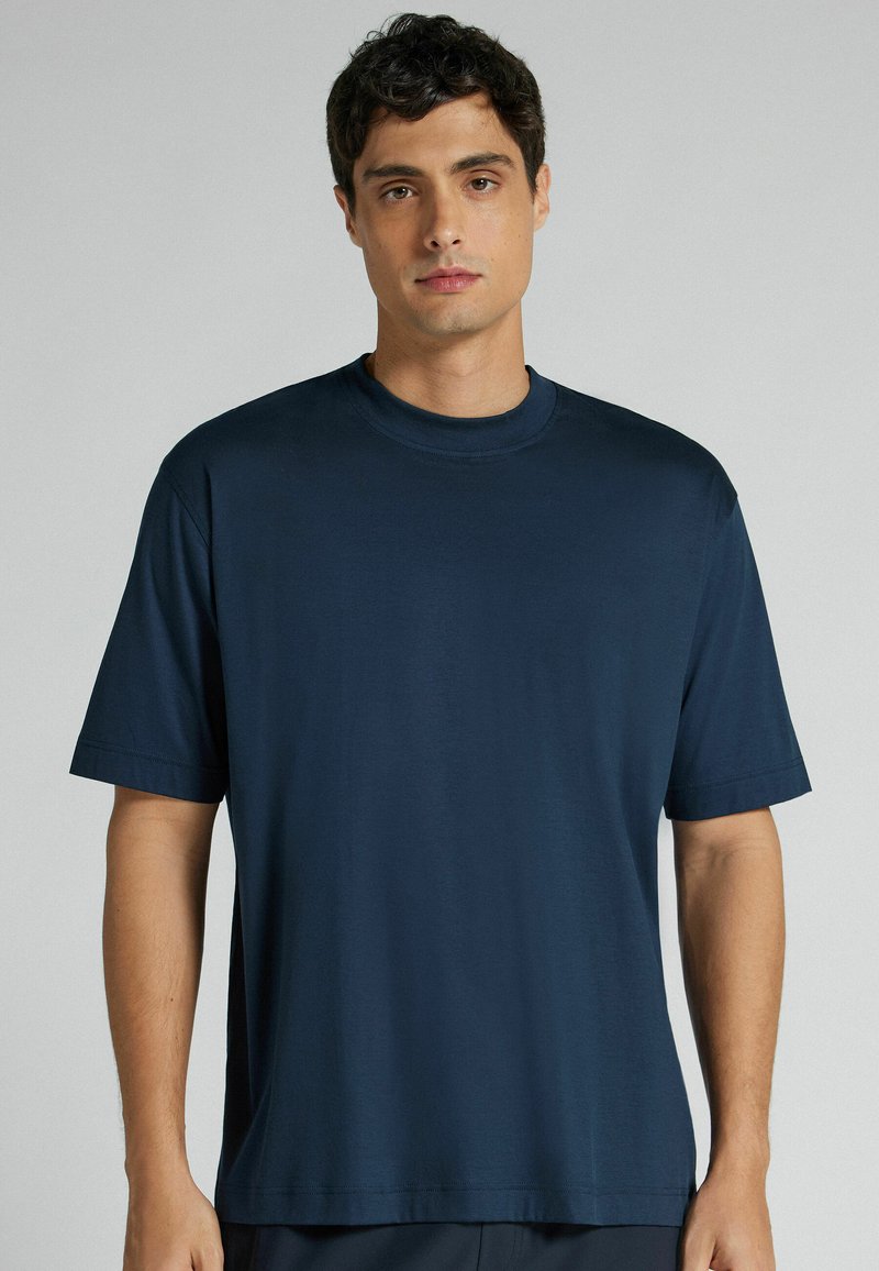 Mann mit kurzen dunklen Haaren, der ein schlichtes dunkelblaues T-Shirt mit kurzen Ärmeln trägt, steht vor einem neutral grauen Hintergrund.