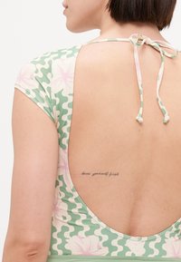 Maillot de bain vert et crème avec des motifs floraux, dos nu et un détail de nœud au cou ; présente un tatouage visible.