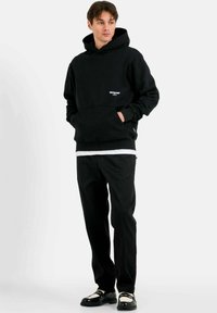 Felpa nera con tasca a marsupio e logo bianco, abbinata a pantaloni neri e scarpe con accenti bianchi, su uno sfondo neutro.