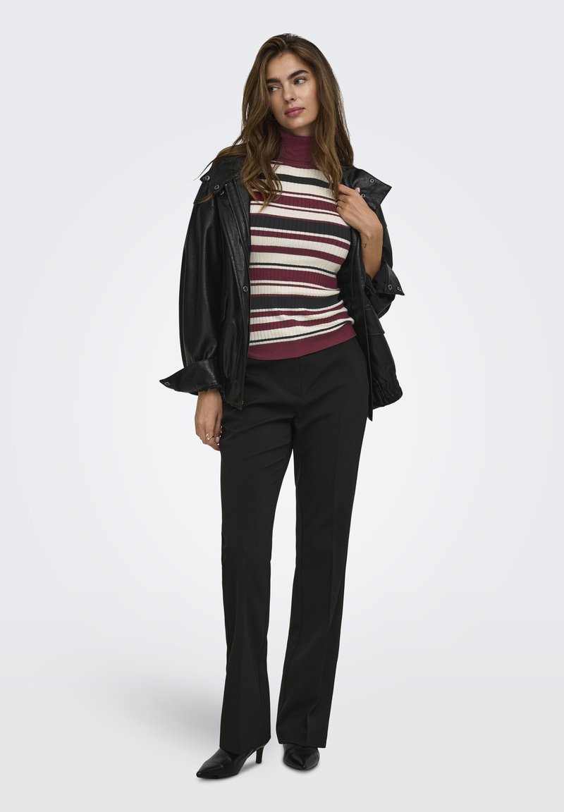 Femme portant une veste en cuir noire sur un pull à col roulé rayé bordeaux et blanc, un pantalon noir et des bottines noires à talons, debout devant un fond gris.
