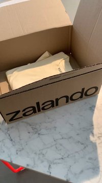 Scatola di cartone marrone con il marchio Zalando, contenente un indumento piegato di colore chiaro, poggiata su una superficie in marmo.