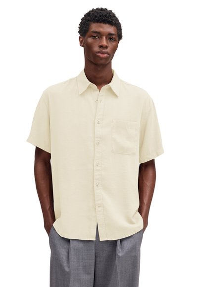 Homme portant une chemise beige à manches courtes avec une poche sur la poitrine et un pantalon gris, debout les mains dans les poches.