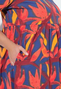 Kirjava kangas, millel on punane, kollane ja sinine lillemuster; omab kogutud taljet ja küljetaskut, mis on kujundusse integreeritud.