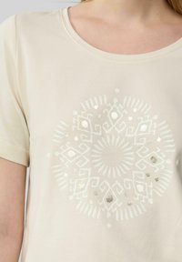 T-shirt beige clair à manches courtes, avec un motif mandala métallique argenté central, texture lisse et col rond.