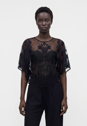 Femme portant un haut en dentelle noire transparente avec motifs floraux et un pantalon noir à cordon, debout devant un fond blanc uni.