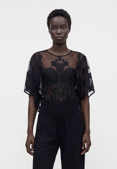 Femme portant un haut en dentelle noire transparente avec motifs floraux et un pantalon noir à cordon, debout devant un fond blanc uni.