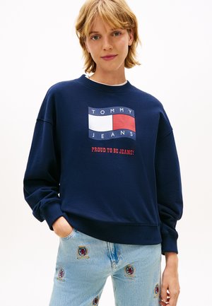 Mladá osoba s krátkymi blond vlasmi má na sebe tmavomodrú mikinu s logom „Tommy Jeans“ a svetlomodré džínsy s vyšívanými záplatami.