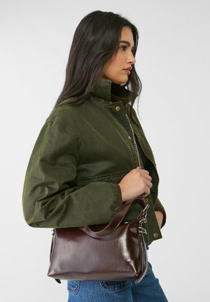 Braune Lederhandtasche mit glatter Textur, runder Form, oberen Griff und silbernen Beschlägen, an einer grünen Jacke gehalten.