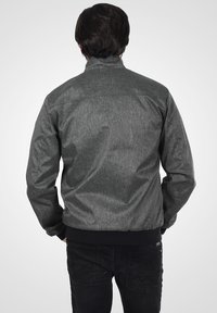 Grå teksturert jakke med ståkrage, elastiske mansjetter og svart ribbestrikket midje. Designet er uformelt og tettsittende.