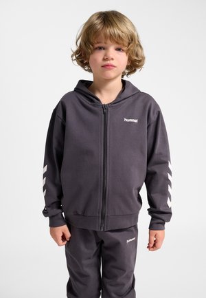 Dziecko z blond włosami ubrane w ciemnoszarą bluzę z kapturem na zamek oraz pasujące spodnie z białymi paskami w kształcie litery V na rękawach, stojące na jednolitym tle.