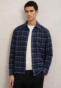 Camicia a quadri blu e nera in tessuto morbido, con bottoni frontali e una tasca sul petto, indossata sopra un maglione bianco strutturato.