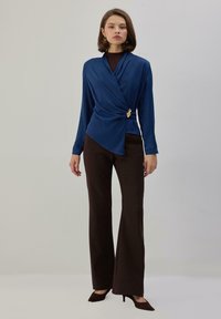 Top azul envolvente con mangas largas y un detalle en forma de hoja dorada en la cintura, combinado con pantalones acampanados marrones y tacones marrones. Tela lisa.