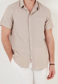 Camicia beige a maniche corte con bottoni in un tessuto leggero, dotata di taschino sul petto e texture morbida, completata da bottoni bianchi.