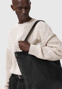 Čierna kožená taška na rameno s textúrovaným povrchom, veľkého obdĺžnikového tvaru a hladkým uchom, ktorú drží osoba oblečená do krémového svetra s pleteným vzorom.