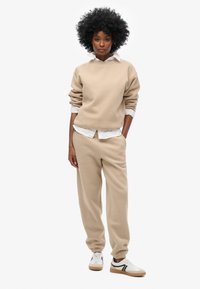 Conjunto de sudadera y pantalones de chándal beige, hechos de tejido suave. Ajuste relajado, puños acanalados y detalle del logo en los pantalones. Camiseta blanca debajo.