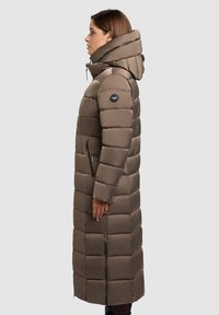 Ilgas pūkuotas paltas taupe spalvos su siuvinėtu raštu, aukštu apykakliu ir užtrauktuko detalėmis šonuose. Kairėje rankovėje yra logotipo pleistras.