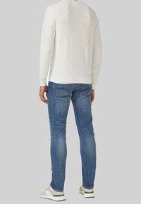 Man die een licht beige trui met lange mouwen, blauwe slim-fit jeans en witte sneakers draagt, staande tegen een effen grijze achtergrond.
